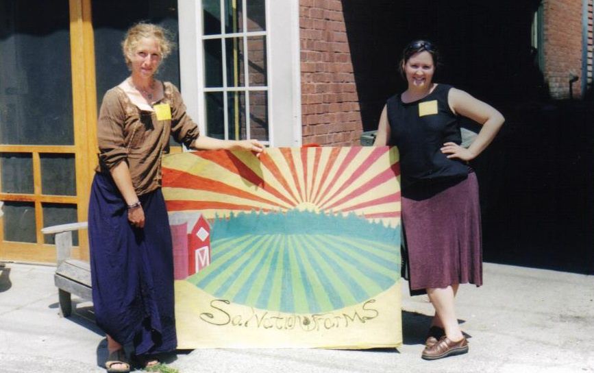 The S.F. Girls two women stand next to a painted sign that reads Salvation Farms. There is a red barn and green fields in the background of the sign